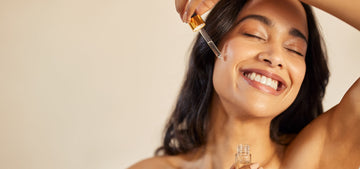 Smiling woman applying a clear serum drop to her cheek with a glass dropper