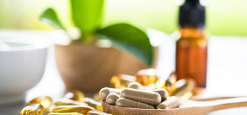 Wooden spoon with tan capsules beside golden gel capsules, an amber dropper bottle, and a green plant