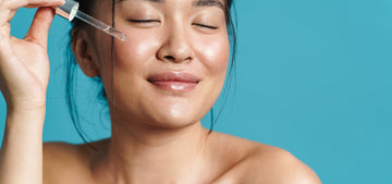 Woman with eyes closed holding a glass dropper with a drop of serum near her cheek